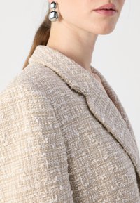 Beige tekstuuri blazer, millel on lõikega krae. Kanga pinnal on kudumise muster, millel on valged aktsendid ja pehme, veidi kõrgendatud pind.