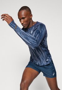 Camisola atlética de manga longa na cor azul-marinho com um padrão de riscas azuis. Apresenta um decote arredondado e tecido elástico. Combinada com calções azuis-marinho.