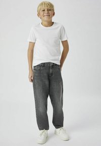 Pojke som bär en vit bomullströja och löst sittande grå denimjeans med knappstängning. Sneakers är vita med en texturerad sula.