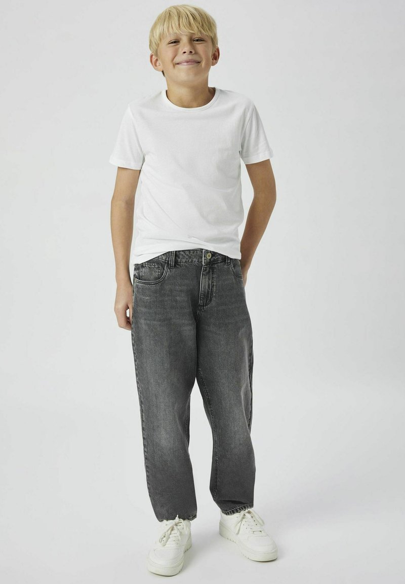 Pojke som bär en vit bomullströja och löst sittande grå denimjeans med knappstängning. Sneakers är vita med en texturerad sula.
