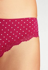 Culotte rouge à pois avec une finition en dentelle, offrant une texture lisse et une taille élastiquée pour plus de confort. Le design comprend des motifs circulaires blancs.