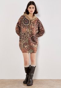 Vestido de sudadera con capucha en tonos marrones con patrones circulares intrincados. Textura suave, corte holgado y detalle de bolsillo frontal. Combinado con botas altas negras.