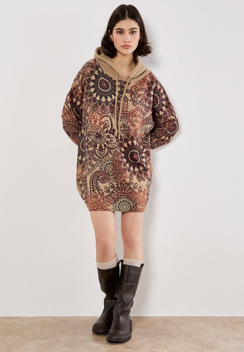 Vestido de sudadera con capucha en tonos marrones con patrones circulares intrincados. Textura suave, corte holgado y detalle de bolsillo frontal. Combinado con botas altas negras.