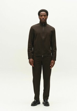 Mann steht frontal, trägt eine dunkelbraune Jacke mit Reißverschluss, passende Hose und schwarze polierte Schuhe vor einem schlichten weißen Hintergrund.
