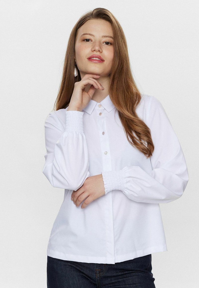 Camicia bianca con maniche lunghe, colletto, chiusura sul davanti con bottoni e polsini arricciati. Tessuto liscio con design minimalista.