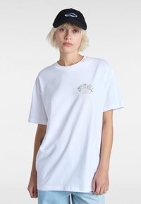 Witte katoenen t-shirt met korte mouwen, met een kleine groene en roze graphic op de borst. Draagt een zwarte baseballpet.