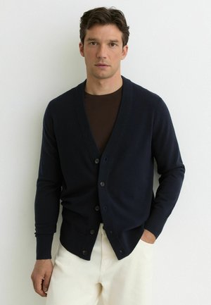 Muž oblečený v tmavomodrom zapínanom kardigane cez hnedú košeľu a biele nohavice, stojaci pred obyčajnou bielou stenou.