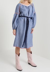 Jeansdress dengan lengan balon, trim merah muda, dan sabuk navy. Dipadukan dengan sepatu cowboy merah muda muda. Tekstur lembut dan potongan santai.
