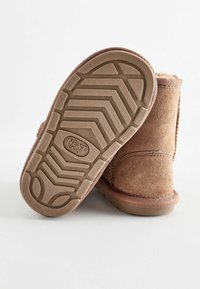 Bruna mockainskor med ankellängd och en texturerad gummisula med ett zigzag-mönster och varumärkeslogotyp, visande bak- och sula vy.