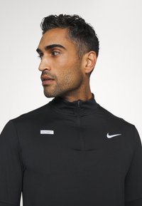 Schwarzes Zip-Up-Athletikoberteil mit Nike-Logo und -Etikett. Sanfte Textur, enganliegendes Design, lange Ärmel und hoher Kragen für Komfort und Leistung.