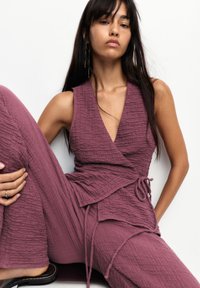 Lila krinklad tyg jumpsuit med omslagdesign, djup V-ringning och knytdekoration vid midjan. Slät textur och byxor med vida ben.