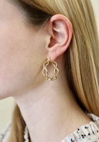Gouden creolen met een golvend ontwerp en een gestructureerd oppervlak, hangend aan de oorlel. Haar is gedeeltelijk zichtbaar, steil gestyled.
