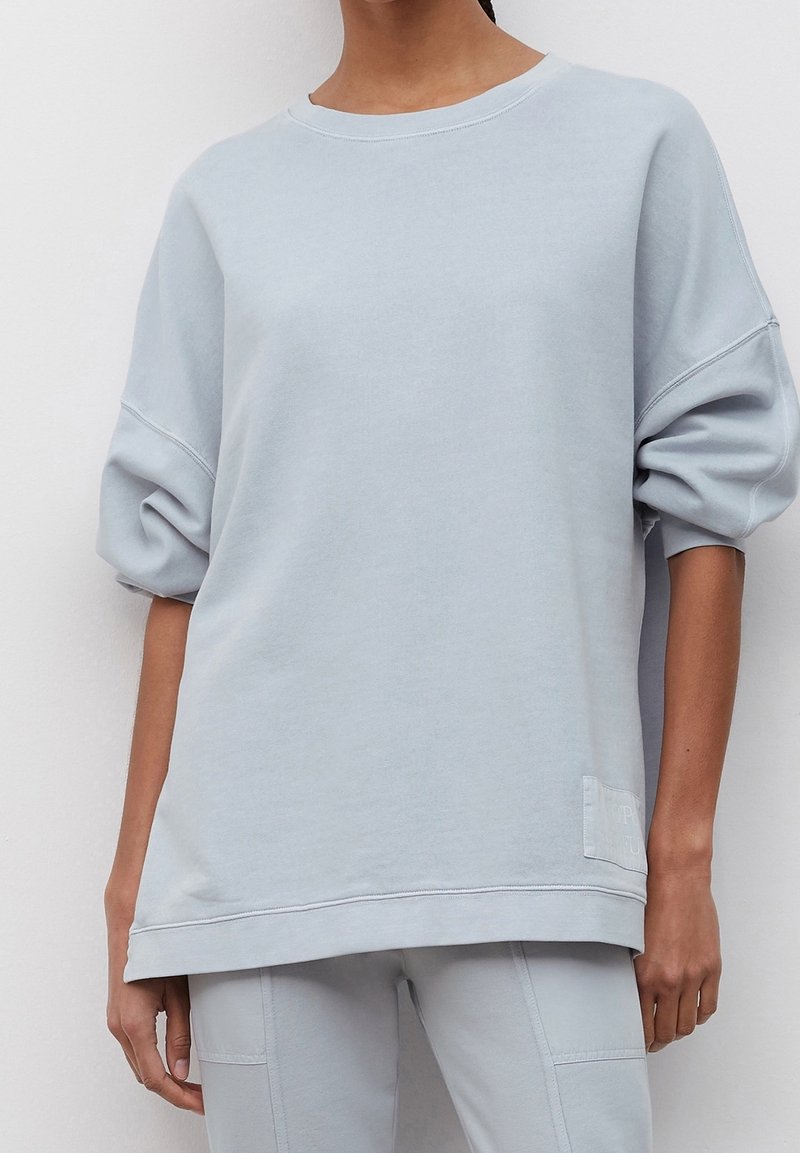 Sweatshirt oversize bleu clair, coton doux, manches larges, col côtelé, détail de patch latéral avec texte, coupe décontractée.