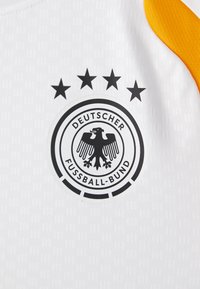 Λευκή αθλητική μπλούζα με πορτοκαλί λεπτομέρειες, φέρει ένα μαύρο λογότ�υπο αετού και πέντε μαύρα αστέρια από πάνω. Κείμενο που γράφει "Γερμανική Ομοσπονδία Ποδοσφαίρου."