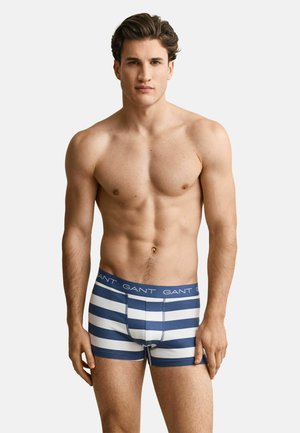 Homme musclé debout de face, portant un boxer bleu et blanc à rayures horizontales avec une ceinture à logo GANT, sur fond blanc.