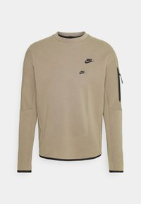 Beige Nike Sweatshirt mit langen Ärmeln, zwei schwarzen Nike-Logos auf der Vorderseite und einer Reißverschlusstasche am linken Ärmel.