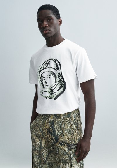 Λευκό βαμβακερό t-shirt με γραφικό αστροναύτη σε μαύρο και πράσινο. Συνδυάζεται με σορτς σε γήινους τόνους με μοτίβα αφηρημένων δέντρων.