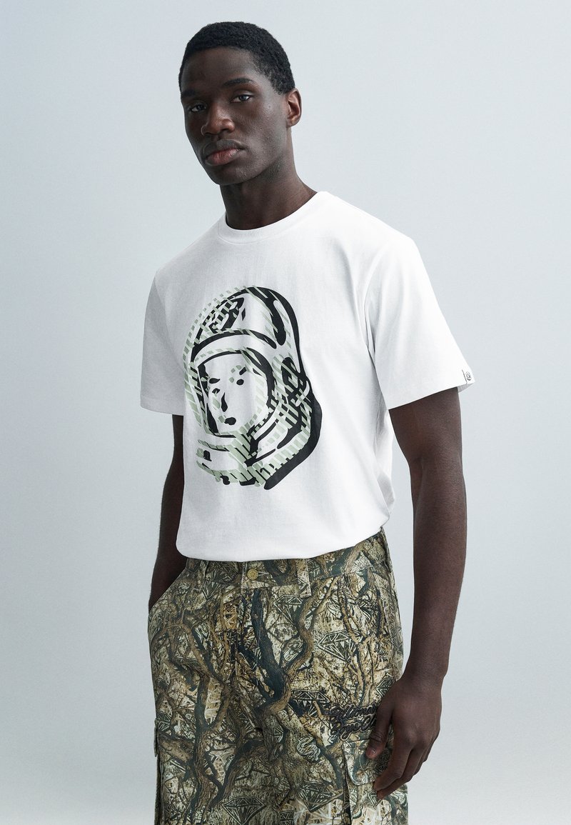 Λευκό βαμβακερό t-shirt με γραφικό αστροναύτη σε μαύρο και πράσινο. Συνδυάζεται με σορτς σε γήινους τόνους με μοτίβα αφηρημένων δέντρων.