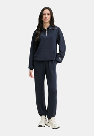 Donna che indossa una felpa blu navy con zip e pantaloni da jogging coordinati con sneakers bianche, in piedi contro uno sfondo chiaro e uniforme.