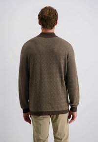 Braun gemusterter Pullover mit strukturiertem Fischgrätmuster, versehen mit einem dunkelbraunen gerippten Kragen und Bündchen, getragen über hellen Hosen.