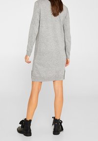 Vestito grigio lavorato a maglia con maniche lunghe e spacchi laterali, abbinato a stivaletti neri con dettagli a nastro. Tessuto liscio, lunghezza al ginocchio.