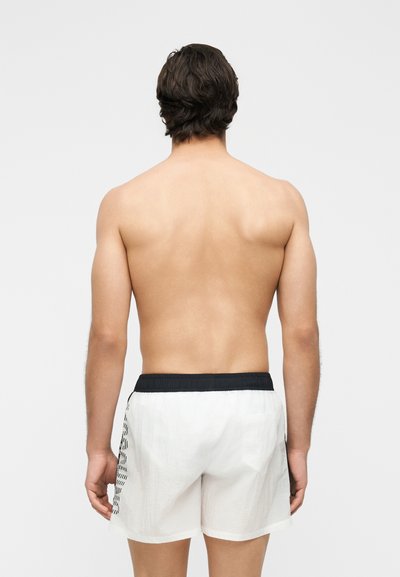 Άνδρας με σκούρα μαλλιά που στέκεται με την πλάτη, χωρίς πουκάμισο, φοράει λευκά σορτς με μαύρη ζώνη και σχέδια στα πλαϊνά, σε απλό φόντο.