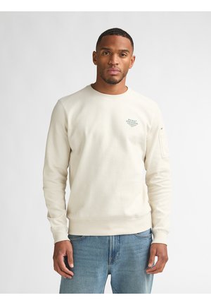 Man draagt een crèmekleurige sweatshirt met lange mouwen en een klein logo op de borst, samen met een blauwe spijkerbroek, staand tegen een effen lichte achtergrond.