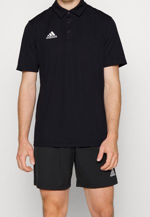 Homme portant un polo noir Adidas et un short noir Adidas, debout devant un fond uni.