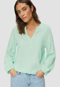Blouse vert menthe avec un col en V, des manches longues et un tissu texturé. Les manches comportent des poignets élastiques et elle est associée à un jean en denim.
