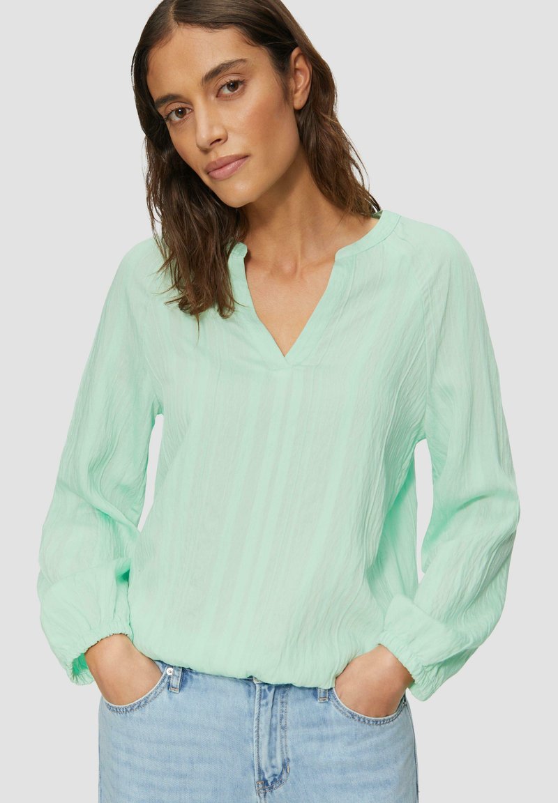 Blouse vert menthe avec un col en V, des manches longues et un tissu texturé. Les manches comportent des poignets élastiques et elle est associée à un jean en denim.