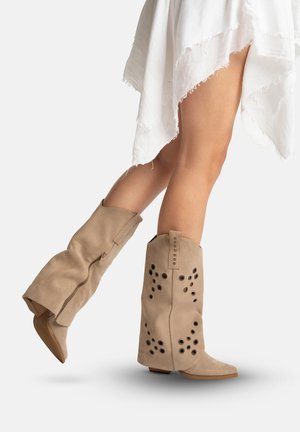 Beine in kniehohen Stiefeln aus beigem Wildleder mit kreisförmigen Ausschnitten, kombiniert mit einem weißen, distressed Rock, die vorwärts auf einem weißen Hintergrund schreiten.