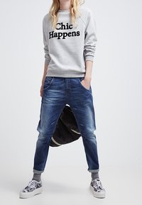 Harmaa huppari, jossa on musta teksti "Chic Happens", yhdistettynä kuluneisiin sinisiin farkkuihin. Mallilla on päällään hopeiset lenkkarit, joissa on paksut valkoiset pohjat.