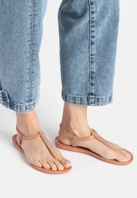 Flache Sandalen in Tan mit einem T-Riemen-Design, verziert mit Kristallakzenten und Nieten, kombiniert mit hellblauen, umgeschlagenen Jeans.