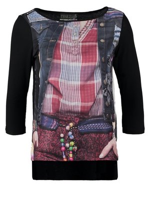 Femme portant une chemise noire à manches longues avec un motif à carreaux imprimé et un design de veste, un jean bleu, des bottes noires, et tenant un sac fourre-tout métallique.