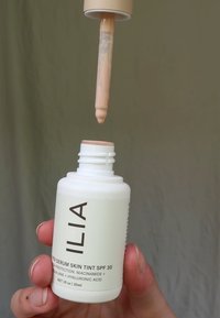 ILIA Super Serum Skin Tint SPF 30 i en vit flaska med svart text, hållande en beige pipett fylld med vätska, mot en neutral bakgrund.