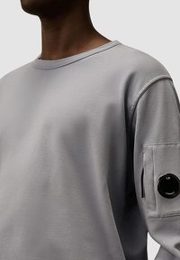 Lichtgrijze langemouwen shirt gemaakt van zachte stof; heeft een ronde halslijn en een zak met ronde hardware detailing op de linkermouw.