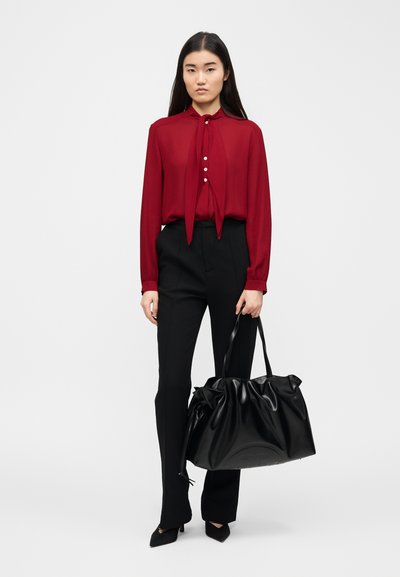 Femme portant un chemisier rouge à manches longues avec un nœud, un pantalon noir, des talons noirs, tenant un grand sac à main noir sur un fond uni.