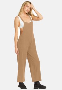 Tan cotton overall with wide legs, deep side pockets, and adjustable shoulder straps. Paired with a white top and black ankle boots.
