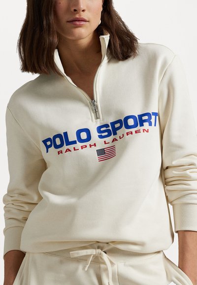 Hanorac crem cu guler cu fermoar pe jumătate, având un logo mare "POLO SPORT" în culori albastre și roșii și un mic detaliu cu steagul american, textură moale.