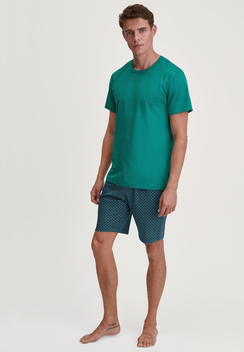Green cotton t-shirt paired with navy swim shorts featuring a wave pattern. Model is barefoot, displaying casual summer attire.