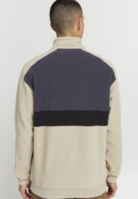 Sudadera de polar en beige con acentos en azul marino y negro. Presenta un cuello alto, corte relajado y tela texturizada. Se muestra la vista trasera.