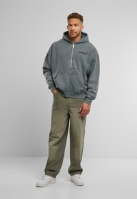 Grå zip-up hoodie med en frontlomme og logo, parret med løstsiddende olivengrønne bukser og hvide sneakers på en grå baggrund.