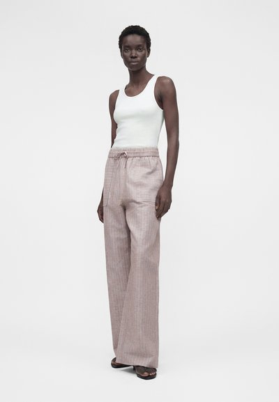 Ψηλή γυναίκα που φοράει λευκό αμάνικο τοπ και άνετο ανοιχτό μπεζ ριγέ παντελόνι με κορδόνι, στέκεται μπροστά σε ένα απλό φωτεινό φόντο.