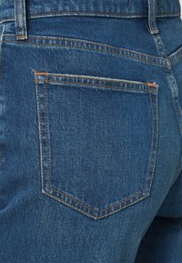 Τζιν με υφή μπλε υφάσματος, που διαθέτει πίσω τσέπη με αντίθετη πορτοκαλί ραφή και κλασικό τετράγωνο σχήμα.