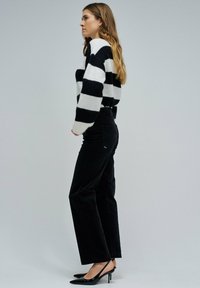 Femme de profil portant un pull à rayures noir et blanc, un pantalon noir taille haute et des escarpins à talons ouverts noirs.