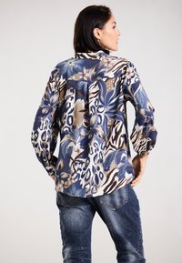 Bluse mit Blumen- und Tierdruck in Blau-, Braun- und Cremetönen. Lange Ärmel, lässiger Schnitt, mit gefälteltem Rücken und Knopfkraagen.