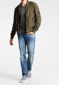 Oliwkowa kurtka typu bomber o krótkim fasonie, z zamkiem z przodu, kieszeniami po bokach i delikatnym połyskiem. Połączona z szarą koszulką, jasnoniebieskimi jeansami i szarymi sneakersami.