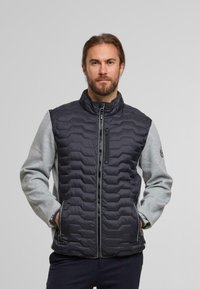 Gilet rembourré avec un devant matelassé noir et des manches en jersey grises. Il dispose d'un col haut, d'une fermeture éclair à l'avant et de poches latérales. Logo sur la manche.