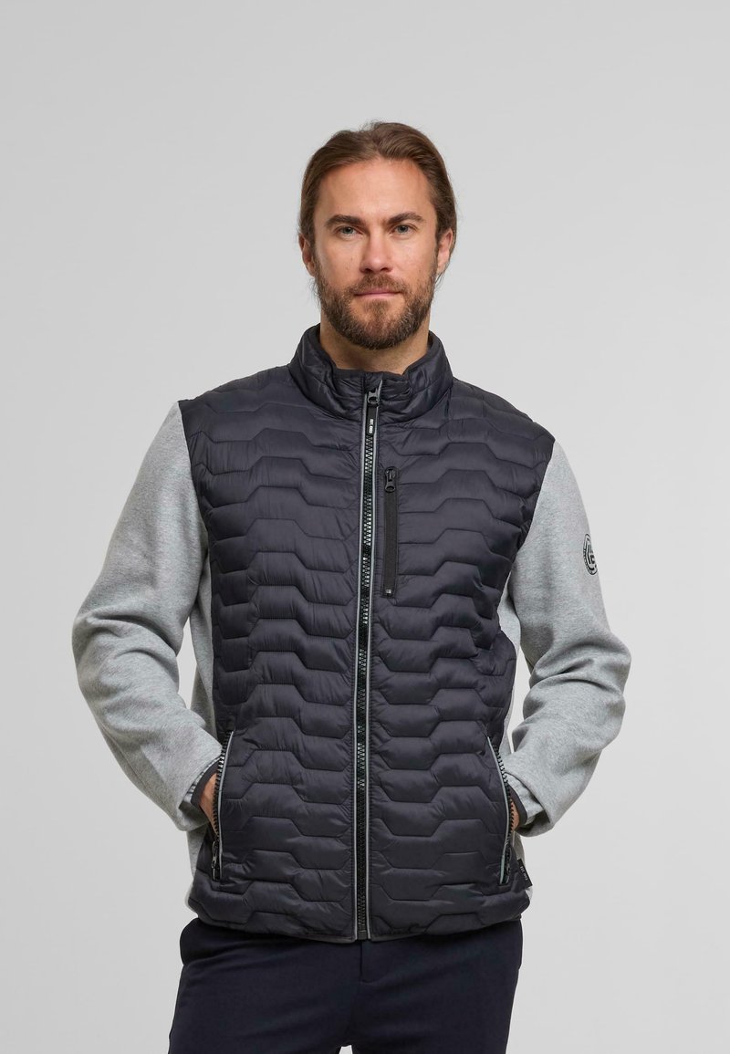 Gilet rembourré avec un devant matelassé noir et des manches en jersey grises. Il dispose d'un col haut, d'une fermeture éclair à l'avant et de poches latérales. Logo sur la manche.