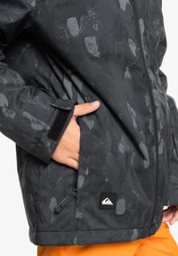 Svart mönstrad jacka med dragkedja, sidoficka med kardborreflik och logotyp patch. Texturerat tyg förbättrar greppet och värmen.